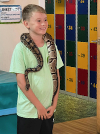 A child standing
            with a python around his neck. A stack of lockers is visible in the background